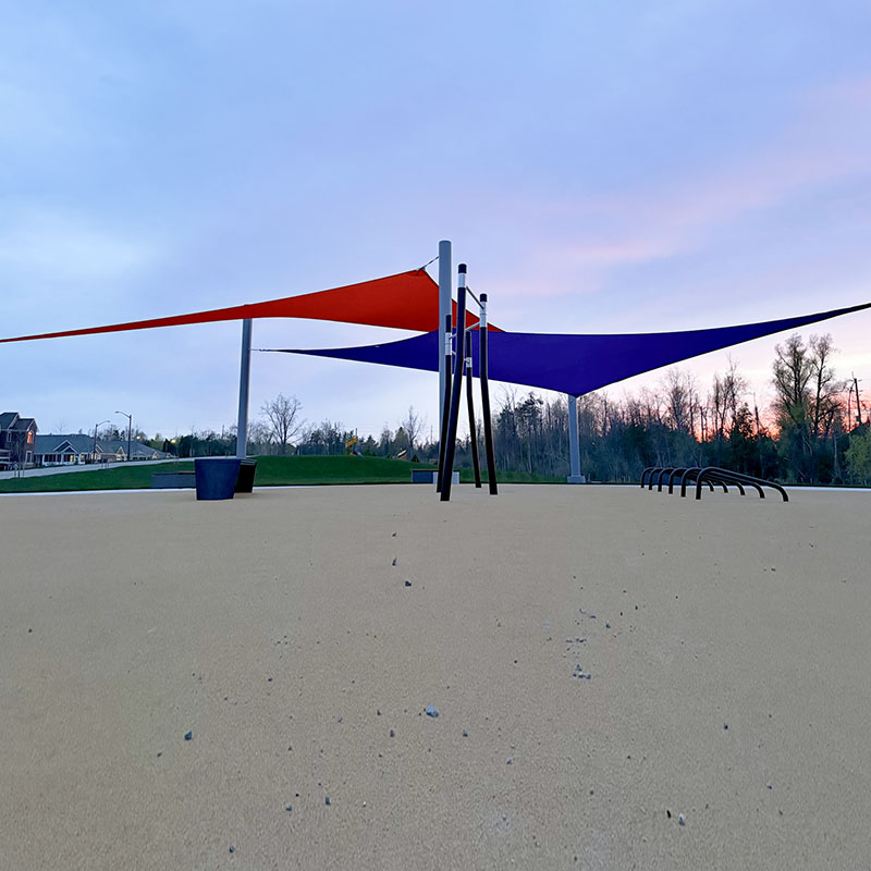 A wide-angle view of a modern outdoor fitness park featuring a dual-layer sun shade system with one orange and one blue triangular sail supported by multiple grey steel posts.