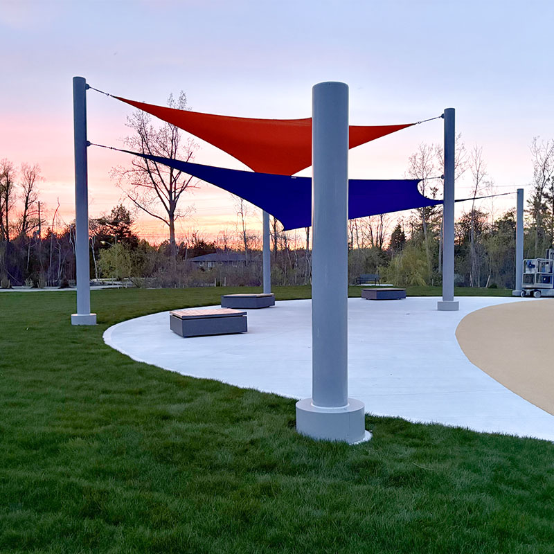 Side profile of a Sun Shade Co. multiple post shade structure at sunset, showing the tensioned cable attachments and two overlapping shade sails in orange and blue providing coverage for a concrete seating area.