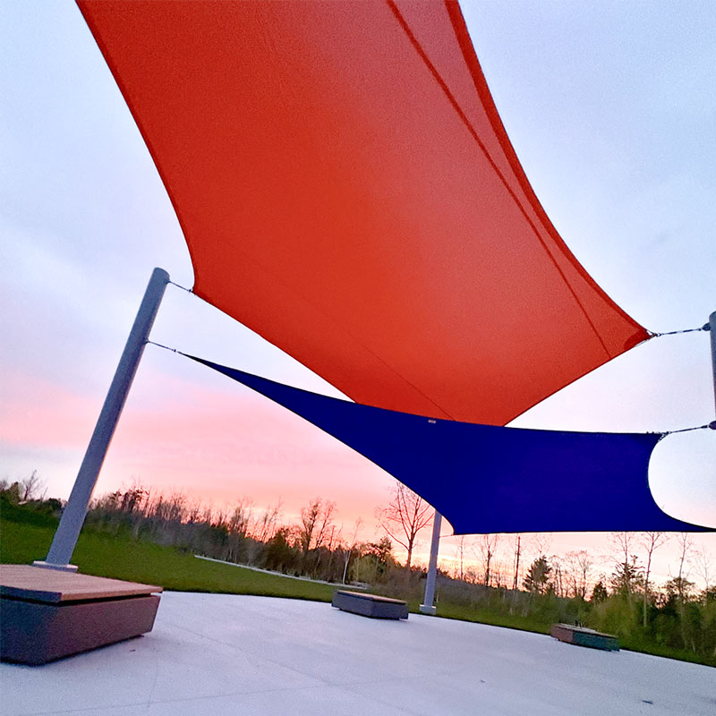 An upward perspective looking through a large orange shade sail and a smaller blue sail, highlighting the heavy-duty steel support posts and the vibrant sky at dusk.