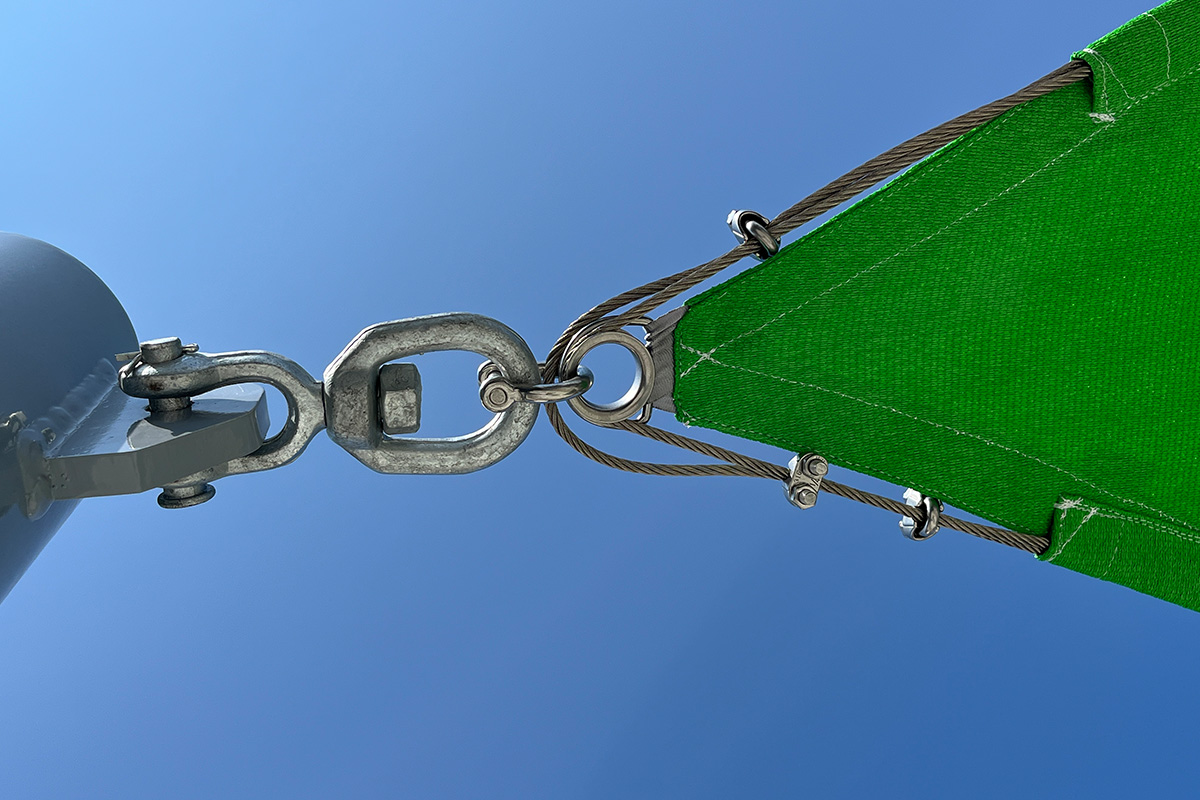 Close-up of heavy-duty stainless steel rigging hardware connecting a bright green commercial shade sail corner to a mounting post.