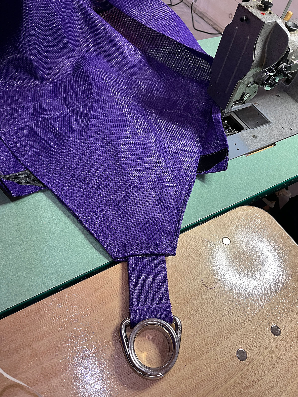 A purple shade sail corner with reinforced webbing and a stainless steel D-ring on a commercial sewing machine table.