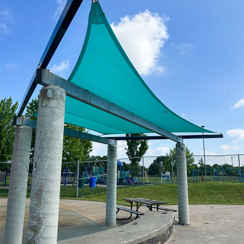 A bright teal commercial shade sail fabric stretched tight over an existing concrete and steel frame structure. A wooden picnic table and concrete bench sit under the canopy, with a green playground visible across a fence under a sunny sky.