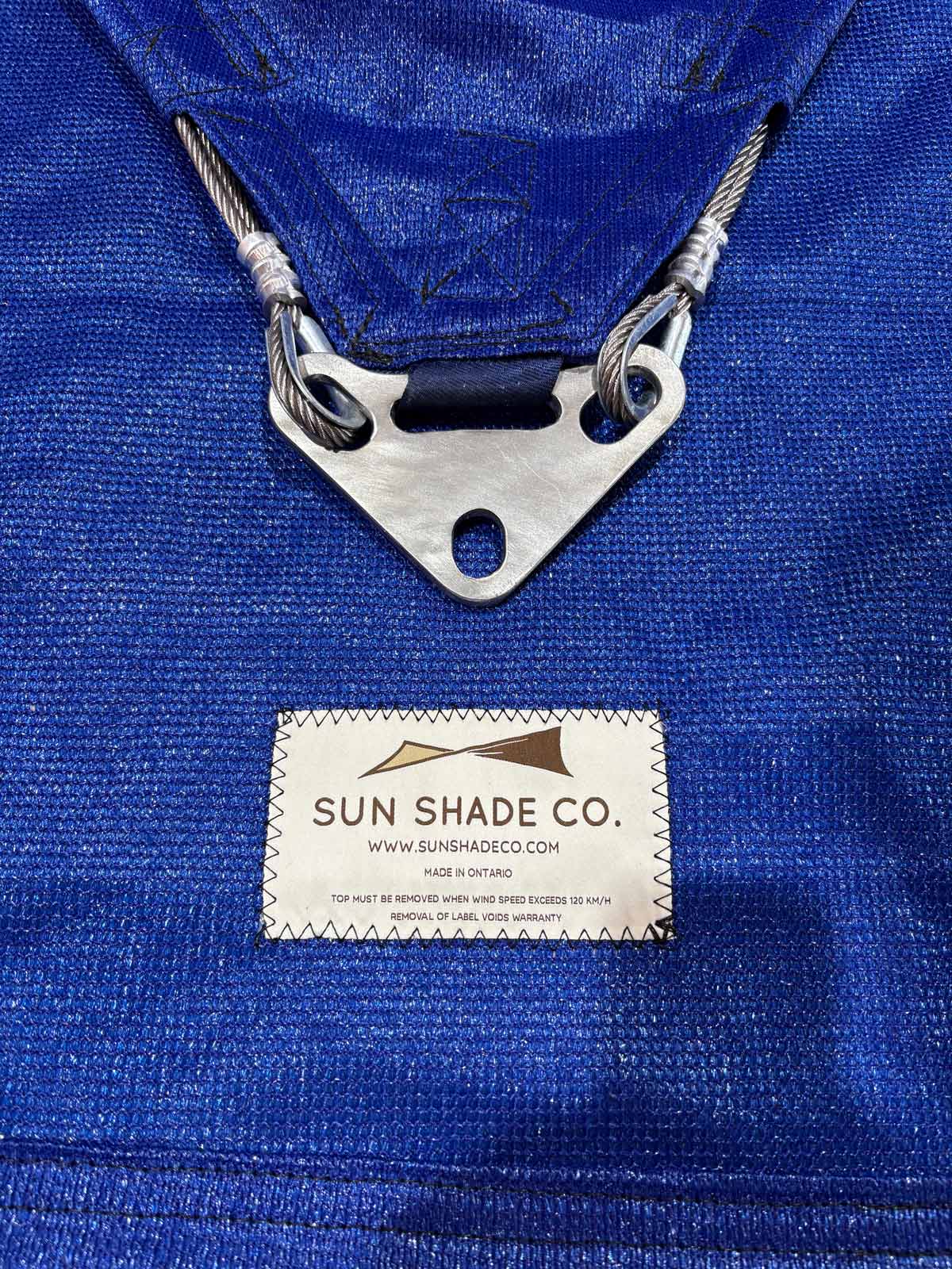 Close-up view of blue fabric shade sail hardware, stainless steel corner plate, and Sun Shade Co. brand label.