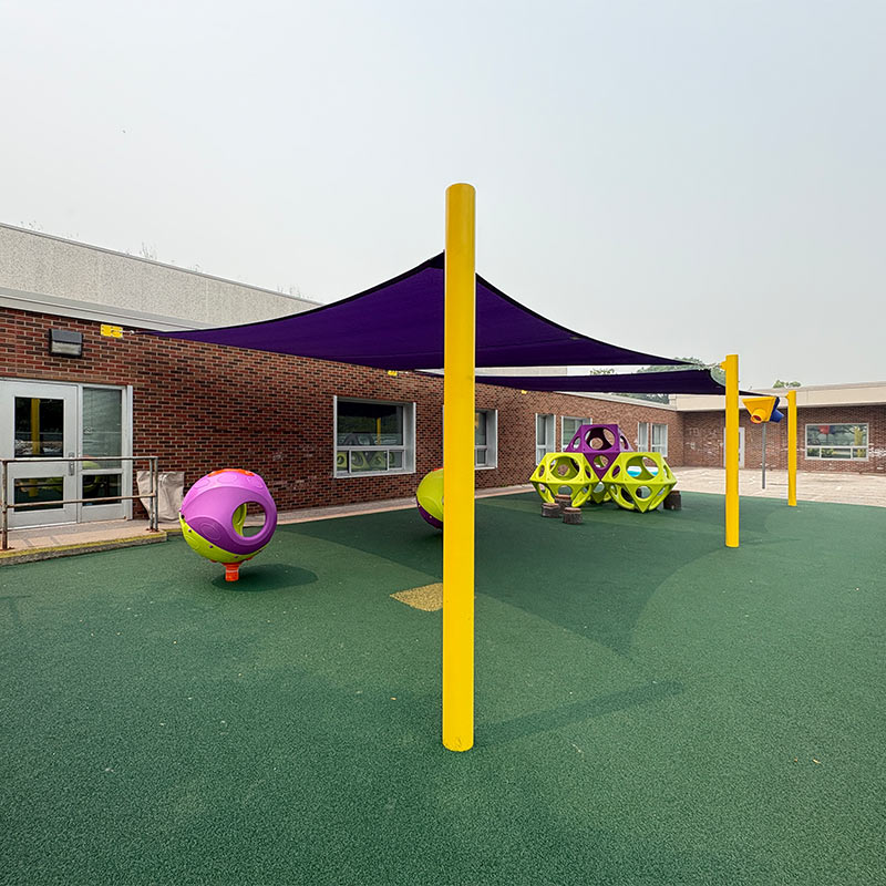 Wide shot showing an installed commercial shade sail system at the Hôtel-Dieu Grace Hospital playground in Windsor. Features Sun Shade Co.’s Super Shade Series with high-density Royal Purple fabric sails attached to wall mounts on the brick building and supported by vibrant Sunbeam Yellow steel posts over a green safety rubber surface.